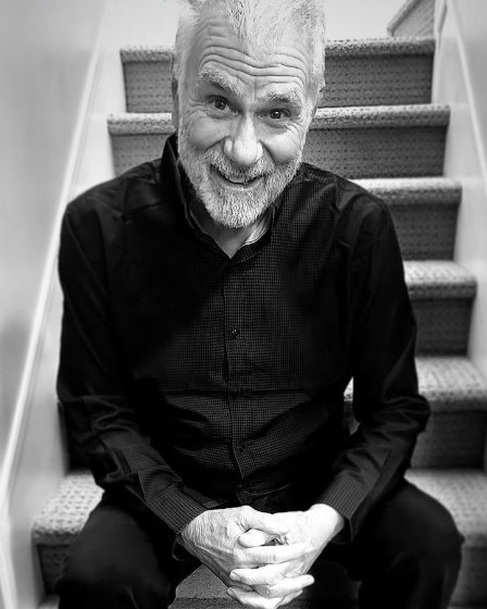 Greig Soohen, seated on a staircase in a black shirt, smiling at the camera.